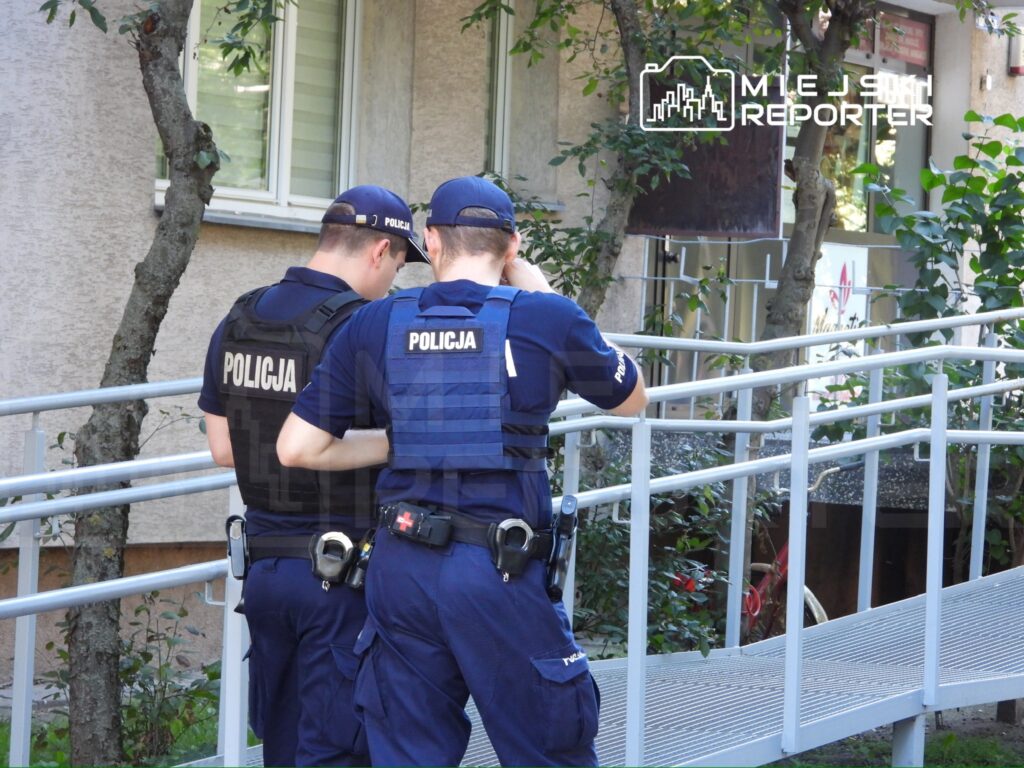 Dwaj policjanci w mundurach stoją na rampie, sprawdzając coś na telefonie w otoczeniu zieleni.