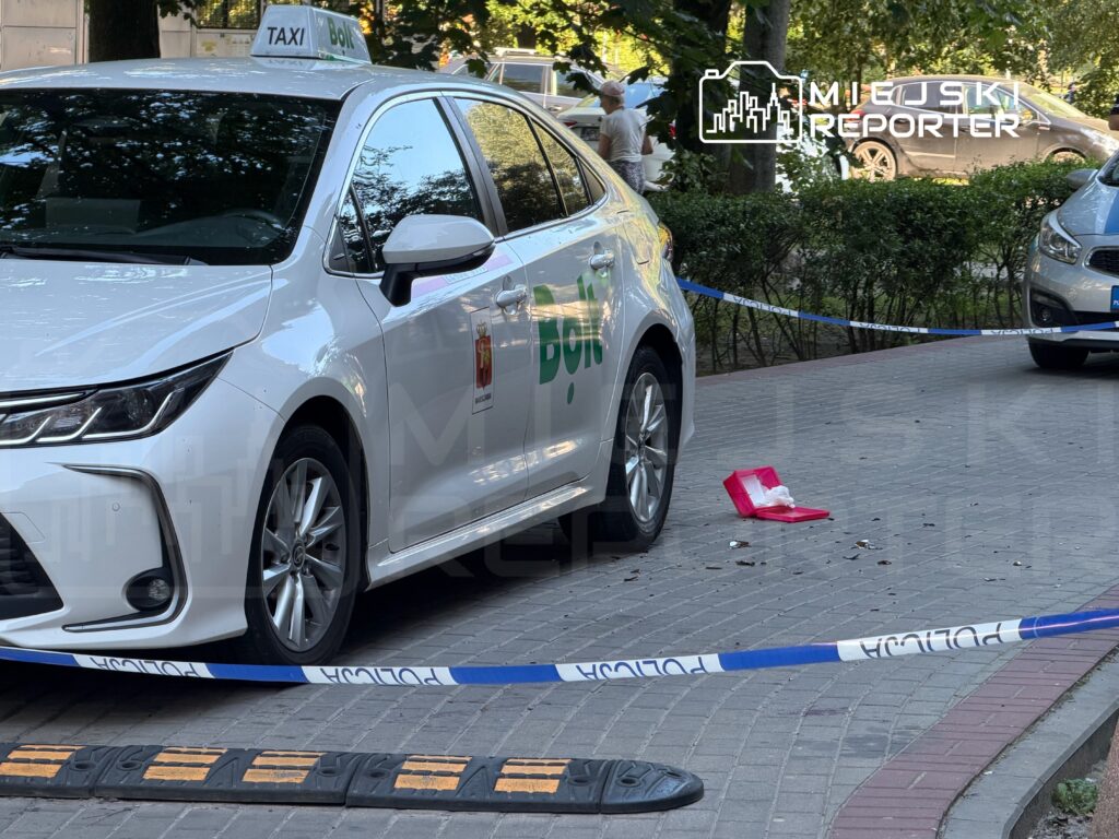 Białe taxi z logo Bolt zaparkowane na chodniku, obok leży różowy pojemnik, a teren otoczony taśmą policyjną.
