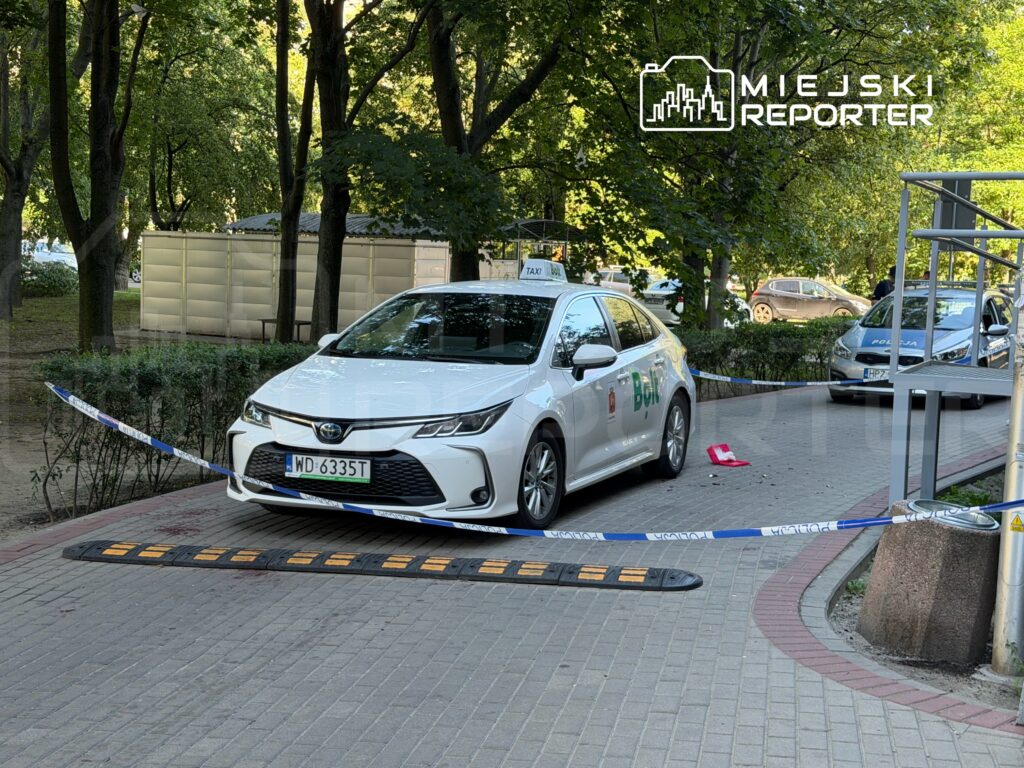 Białe auto osobowe z logo taksówki zaparkowane w otoczeniu zieleni, zabezpieczone taśmą policyjną.