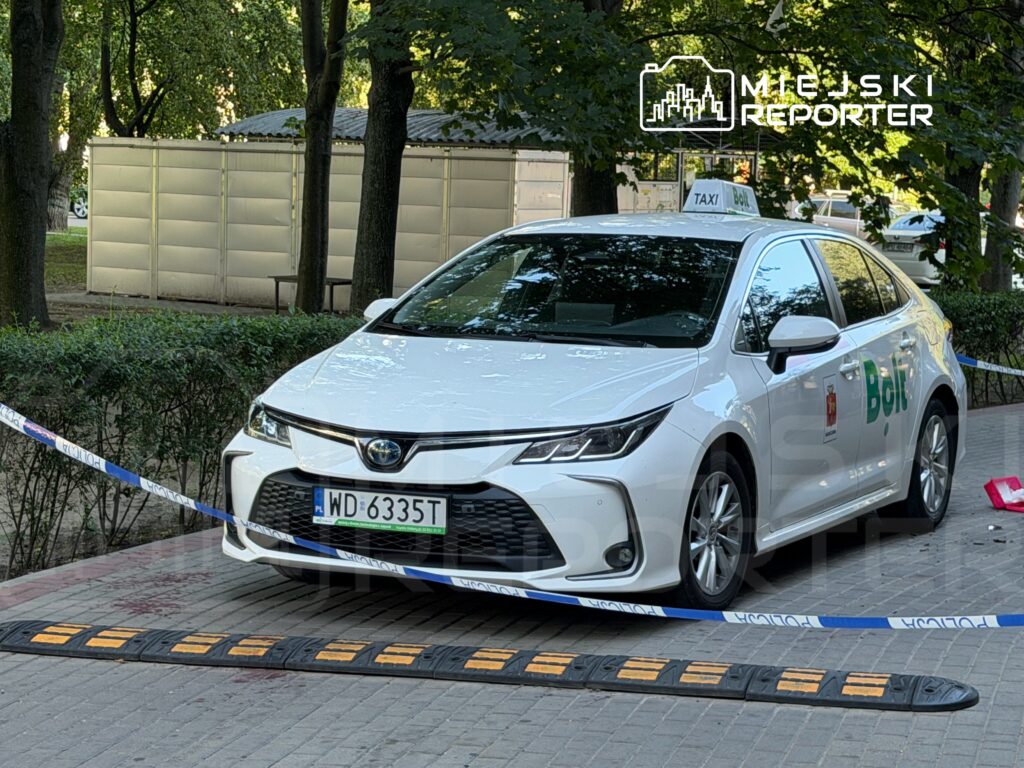 Białe taxi zaparkowane przy chodniku, zabezpieczone taśmą policyjną, w tle zieleń drzew i szare panele.