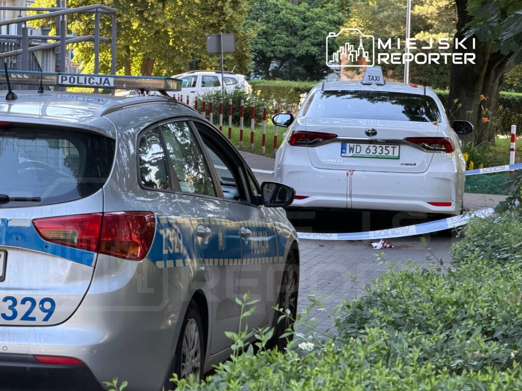 Policyjny radiowóz zaparkowany obok białego taksówki z widocznymi śladami krwi, otoczonego taśmą zabezpieczającą.