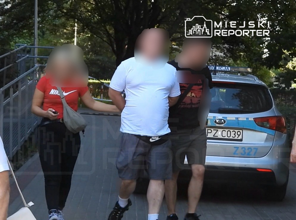 Mężczyzna w białej koszulce prowadzony przez dwóch funkcjonariuszy policji obok radiowozu na tle zieleni.