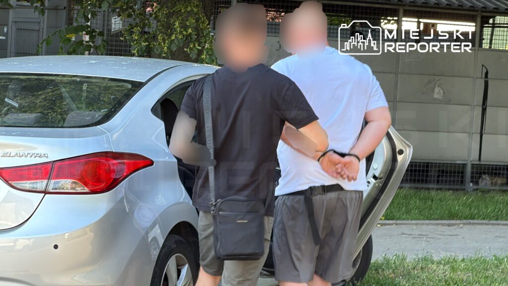 Mężczyzna w białej koszulce z rękami związanymi z tyłu idzie obok mężczyzny w czarnej koszulce, przy samochodzie.
