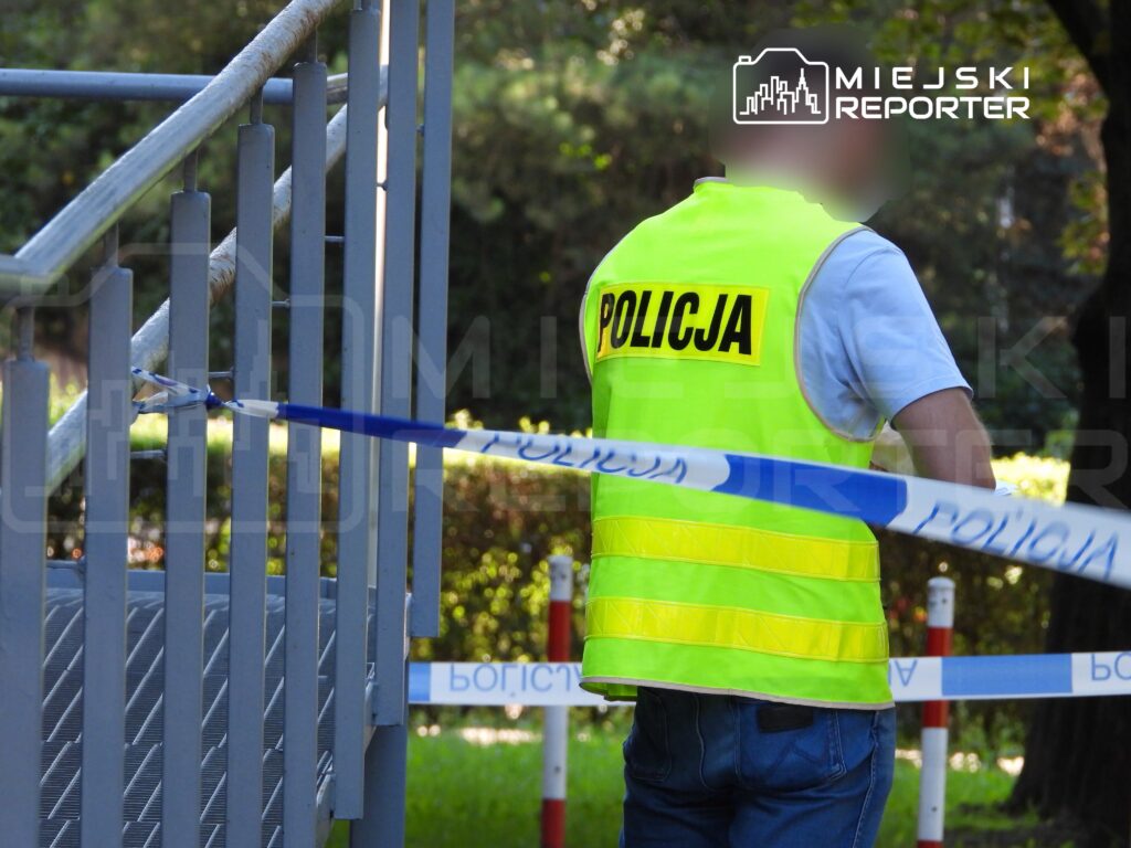 Policjant w żółtej kamizelce zabezpiecza teren oznaczony niebieską taśmą. W tle widać zieleń drzew.