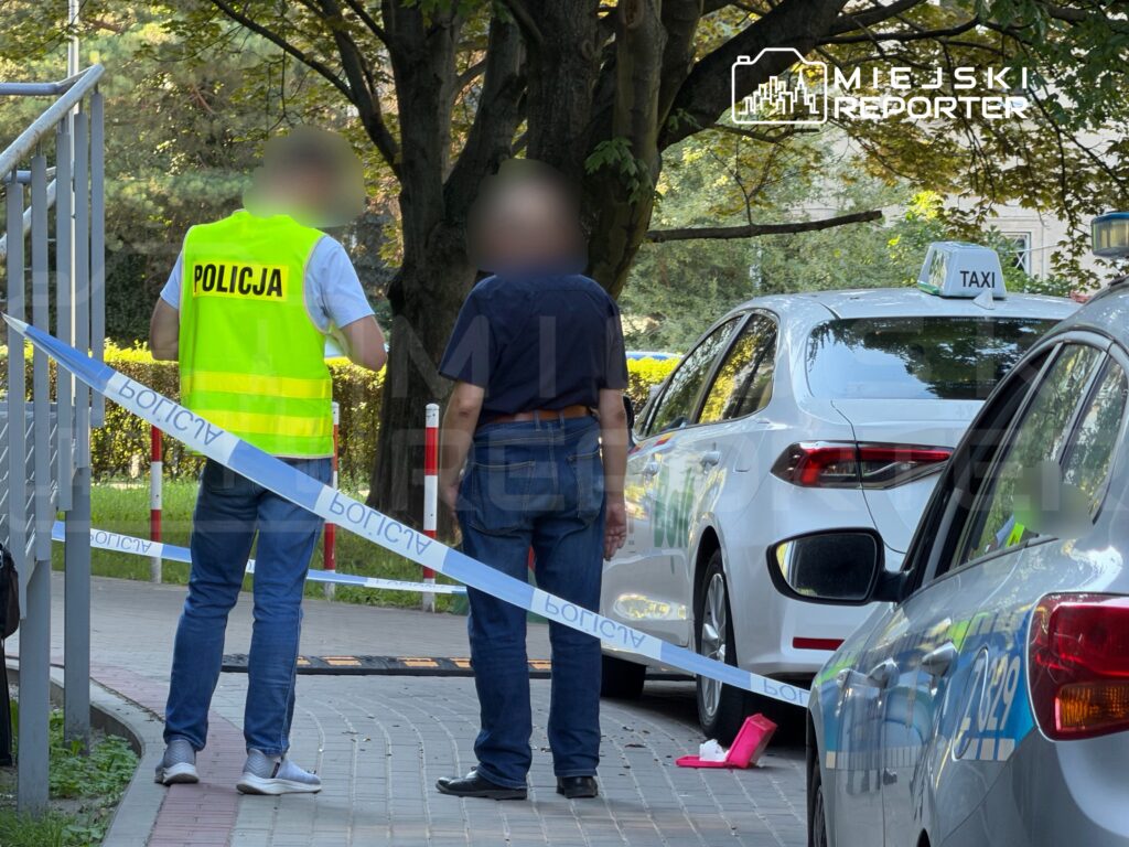 Policjant w kamizelce odblaskowej rozmawia z mężczyzną obok taksówki, na jezdni wyznaczono strefę zabezpieczoną taśmą.
