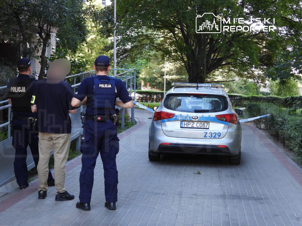 Trzech policjantów prowadzi zatrzymanego mężczyznę wzdłuż chodnika w otoczeniu zieleni, obok radiowozu policyjnego.