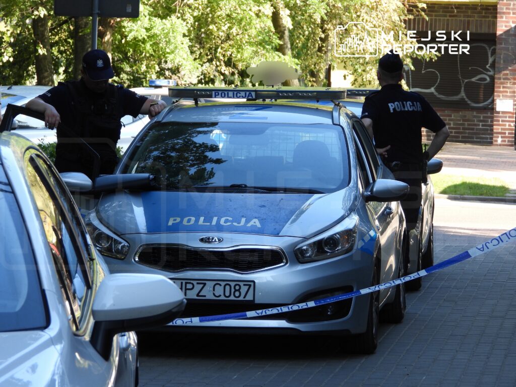 Dwaj policjanci w mundurach stoją obok radiowozu z napisem „POLICJA”, zabezpieczając miejsce zdarzenia.