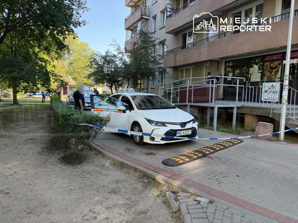 Biały samochód osobowy stoi na chodniku, a obok policjant rozmawia z mężczyzną; w tle widoczna jest budowla z rampą.