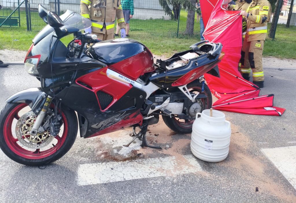 Motocykl w czarno-czerwonym kolorze stoi na drodze, obok widać pojemnik i pracujących strażaków z czerwonym namiotem.