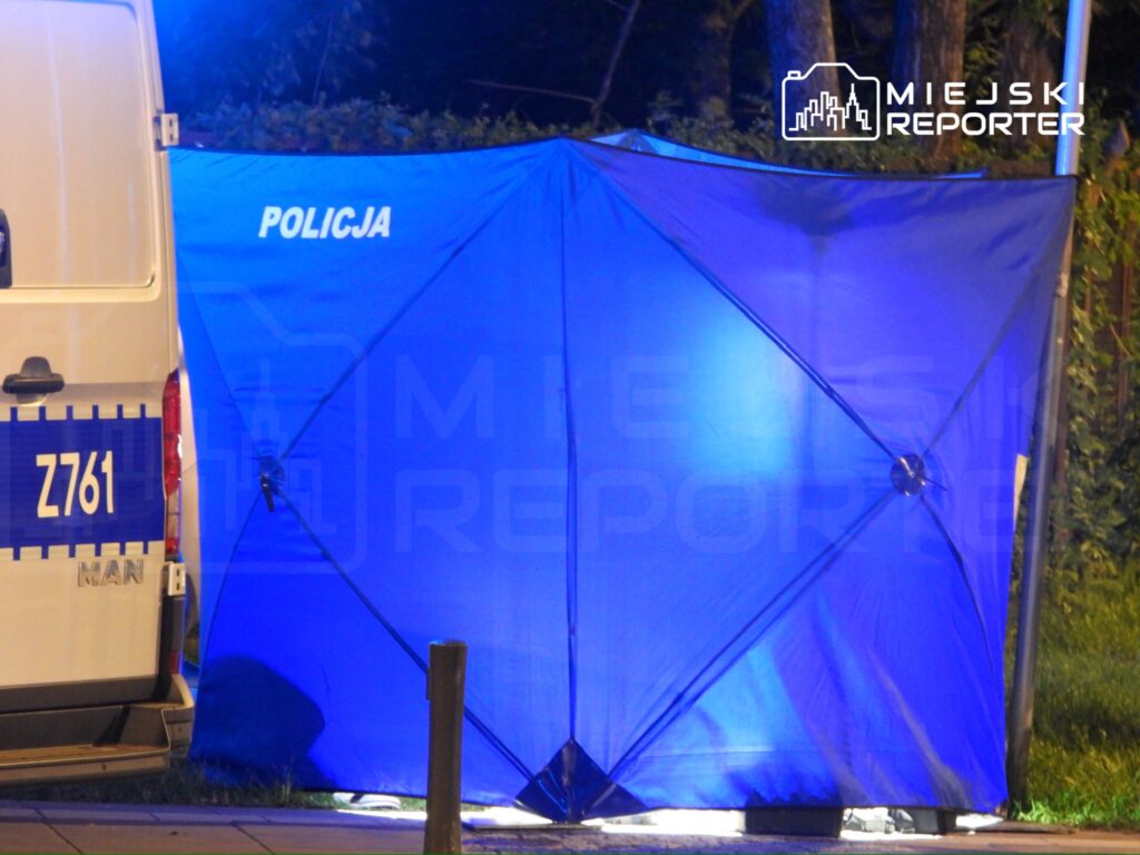 Niebieski namiot policyjny ustawiony obok radiowozu na ulicy, oświetlony przez latarnie.