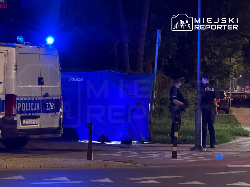 Policjanci rozmawiają obok niebieskiego namiotu na chodniku, obok radiowozu policyjnego.
