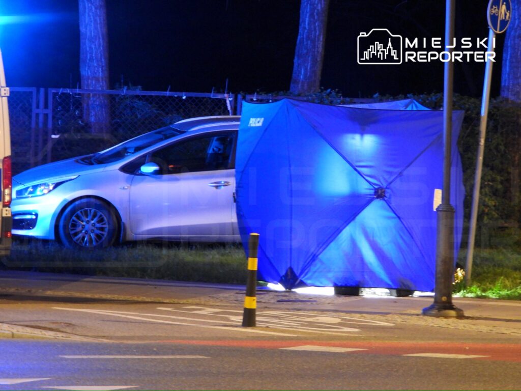 Niebieski namiot policyjny ustawiony obok białego samochodu na oświetlonej ulicy w nocy.
