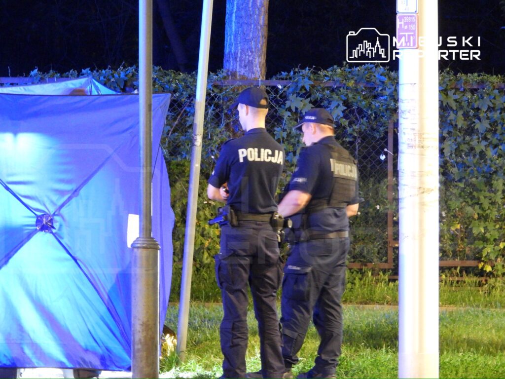 Dwóch policjantów w mundurach rozmawia obok niebieskiego namiotu w ciemnym, zalesionym terenie.