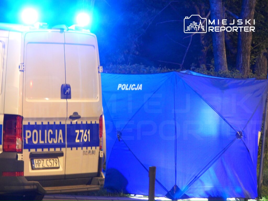 Policyjny van z włączonymi sygnałami świetlnymi stoi obok niebieskiego namiotu z napisem "POLICJA" w ciemnym otoczeniu.