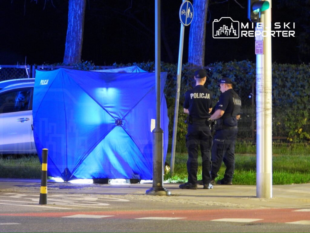 Policjanci rozmawiają obok niebieskiego namiotu na chodniku w nocy, w tle widać samochód i latarnię.