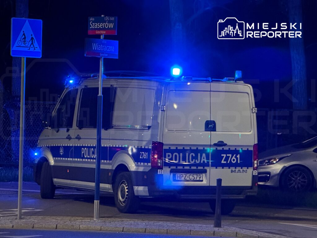 Policyjny furgon z włączonymi światłami na ulicy Szaserów, obok znaków drogowych oraz w otoczeniu ciemnej nocy.