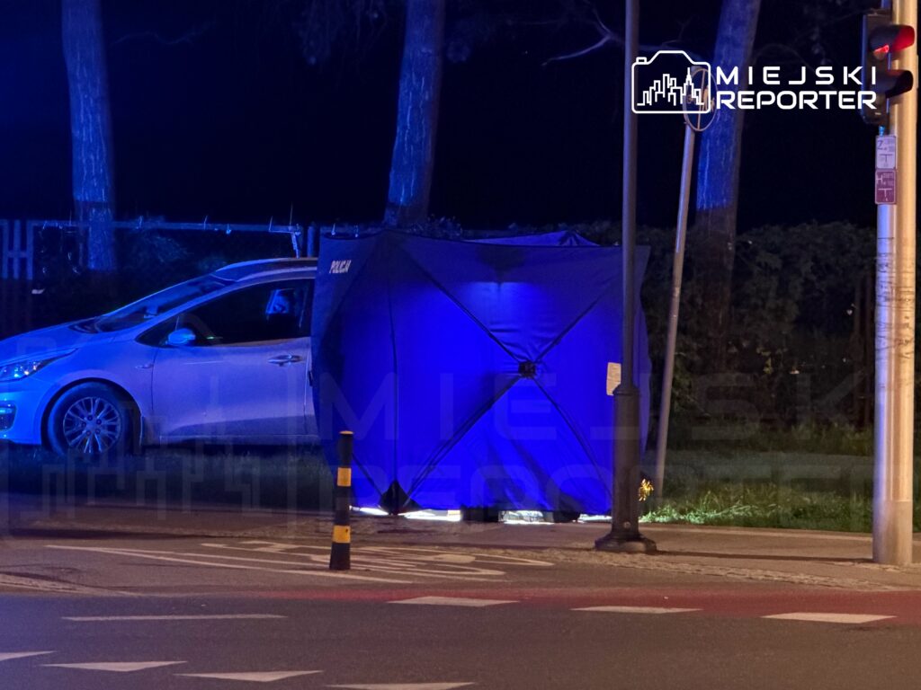 Nocna scena z policyjnym namiotem oświetlonym niebieskimi światłami obok zaparkowanego samochodu.