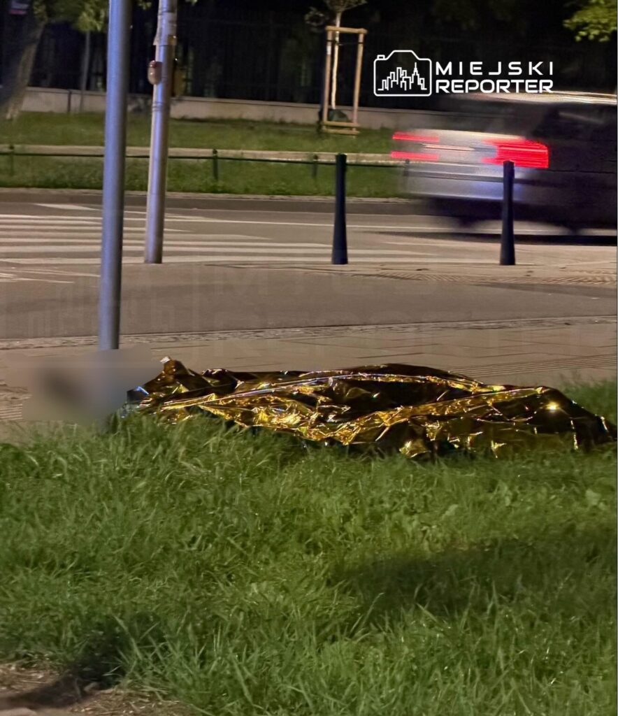 Złota folia ratunkowa leży na trawie obok chodnika w nocy, w tle widać przejeżdżający samochód.