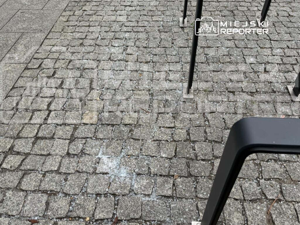 Fragment kostki brukowej z widocznymi plamami, w otoczeniu metalowych słupków.