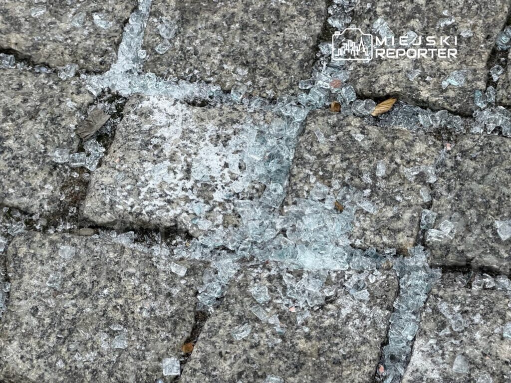 Szkło rozbite na kostce brukowej, wśród drobnych kamieni i liści, tworzy niebezpieczną sytuację na chodniku.