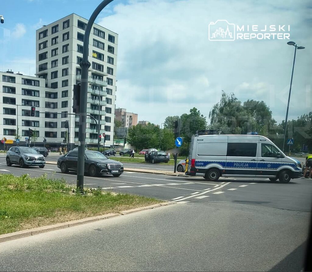 Policyjny radiowóz zatrzymany na rondzie, obok ruchliwych samochodów, w tle nowoczesne budynki mieszkalne i zieleń.