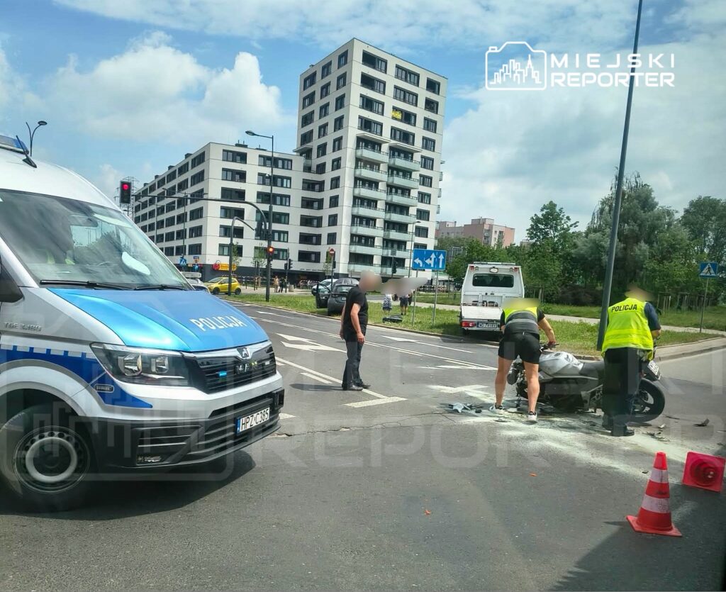 Mężczyzna w garniturze rozmawia z policjantem przy przewróconym motocyklu na skrzyżowaniu w miejskim otoczeniu.