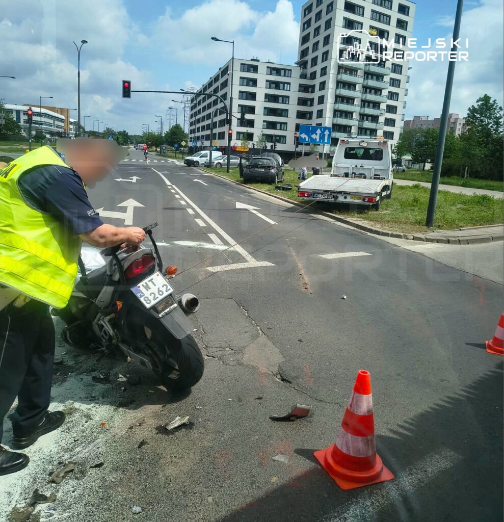 Policjant w kamizelce odblaskowej bada skutki wypadku motocykla na skrzyżowaniu, obok leży uszkodzony pojazd.