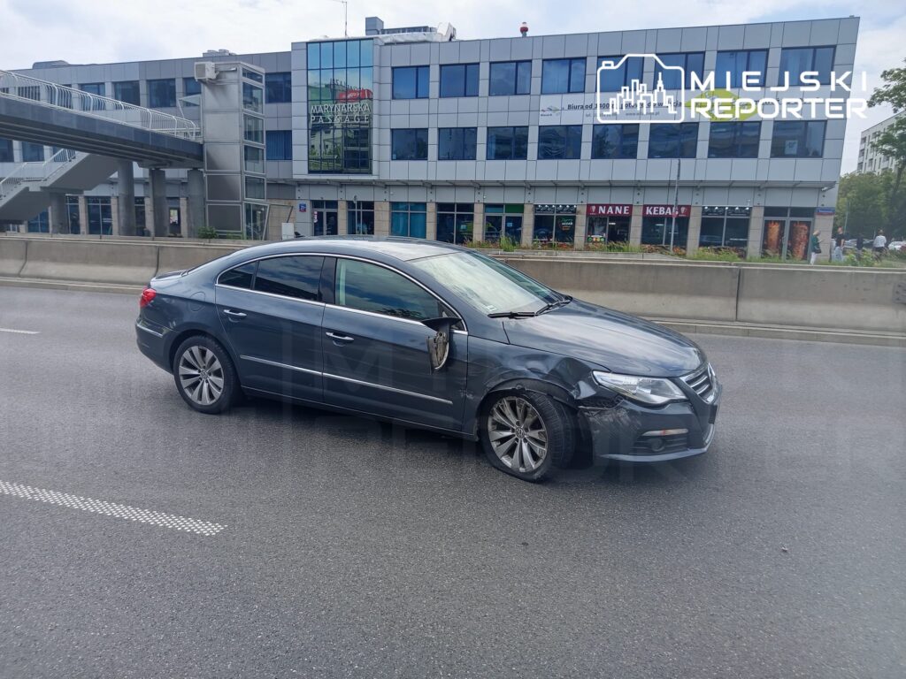 Uszkodzony samochód osobowy stojący na jezdni obok nowoczesnego biurowca z przeszkloną fasadą.