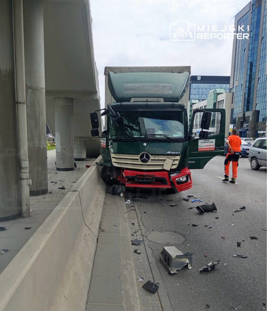 Czerwony i zielony ciężarówka Mercedes z uszkodzoną kabiną stoi przy barierze na ruchliwej ulicy, wokół rozrzucone fragmenty.