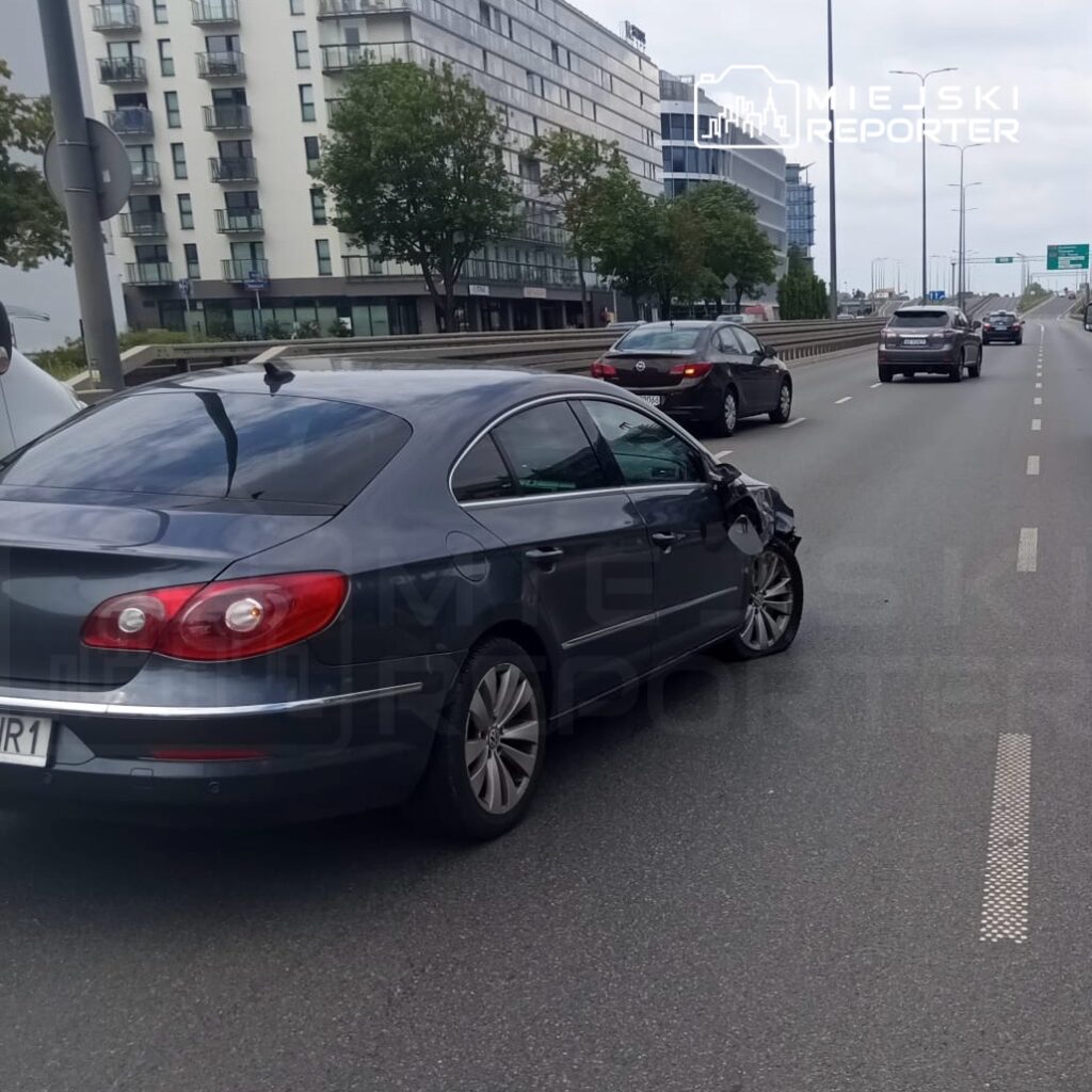 Uszkodzony szary samochód osobowy zaparkowany na pasie ruchu w miejskim otoczeniu, obok innych pojazdów.