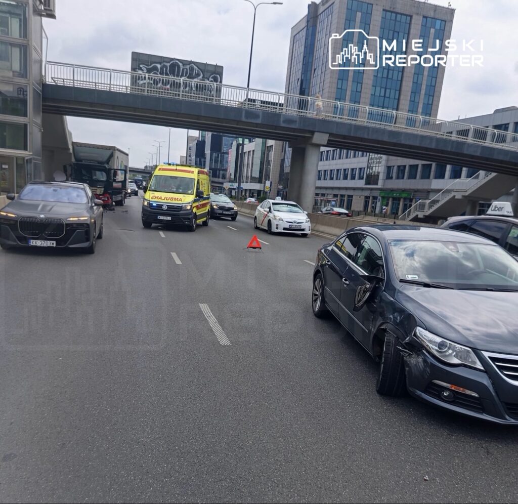 Wielopojazdowy wypadek na ruchliwej ulicy, z ambulansami i uszkodzonymi samochodami oraz trójkątem ostrzegawczym.