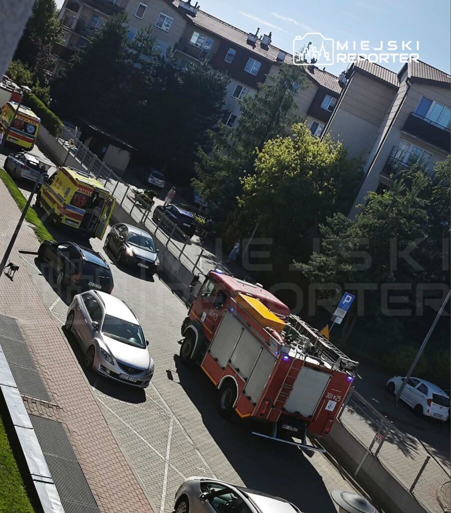 W tle nowoczesne budynki, przed nimi pojazdy ratunkowe, w tym wóz strażacki oraz karetka, zaparkowane przy ulicy.