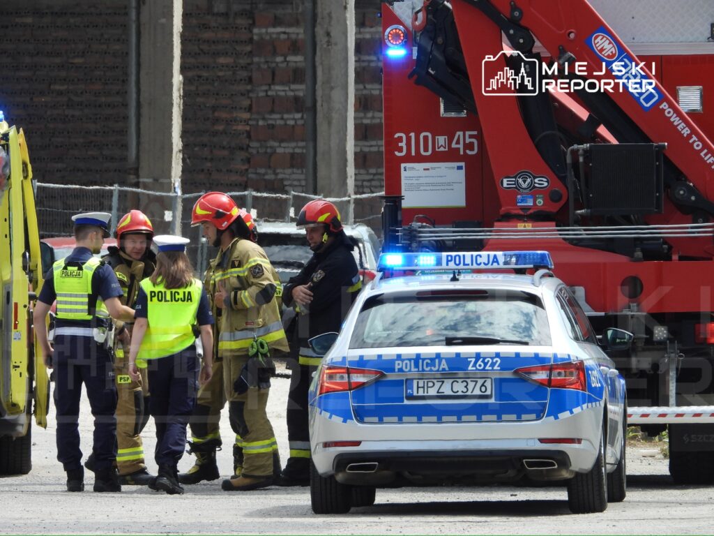 Grupa funkcjonariuszy policji i straży pożarnej rozmawia na placu budowy, obok pojazdu strażackiego i radiowozu.