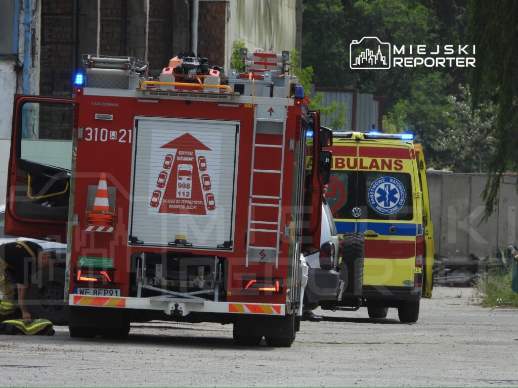 Strażak w mundurze przygotowuje sprzęt obok wozu strażackiego na terenie przemysłowym, a w tle widoczny ambulans.