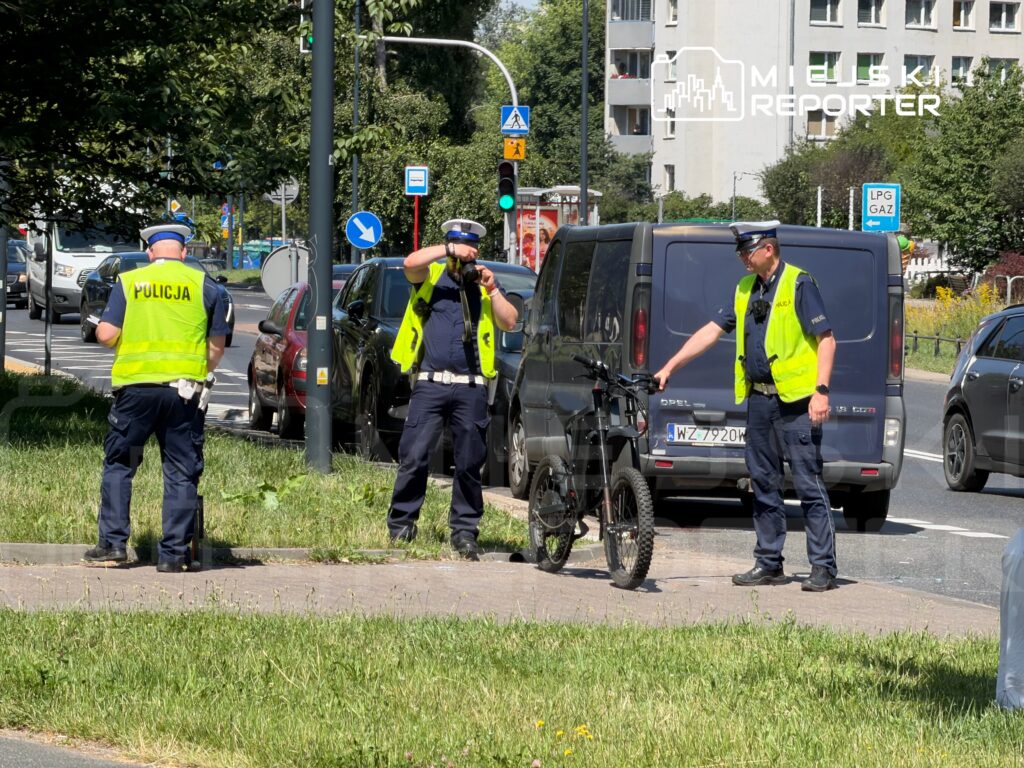 Trzech policjantów w kamizelkach odblaskowych kontroluje rower w pobliżu ruchliwej ulicy.