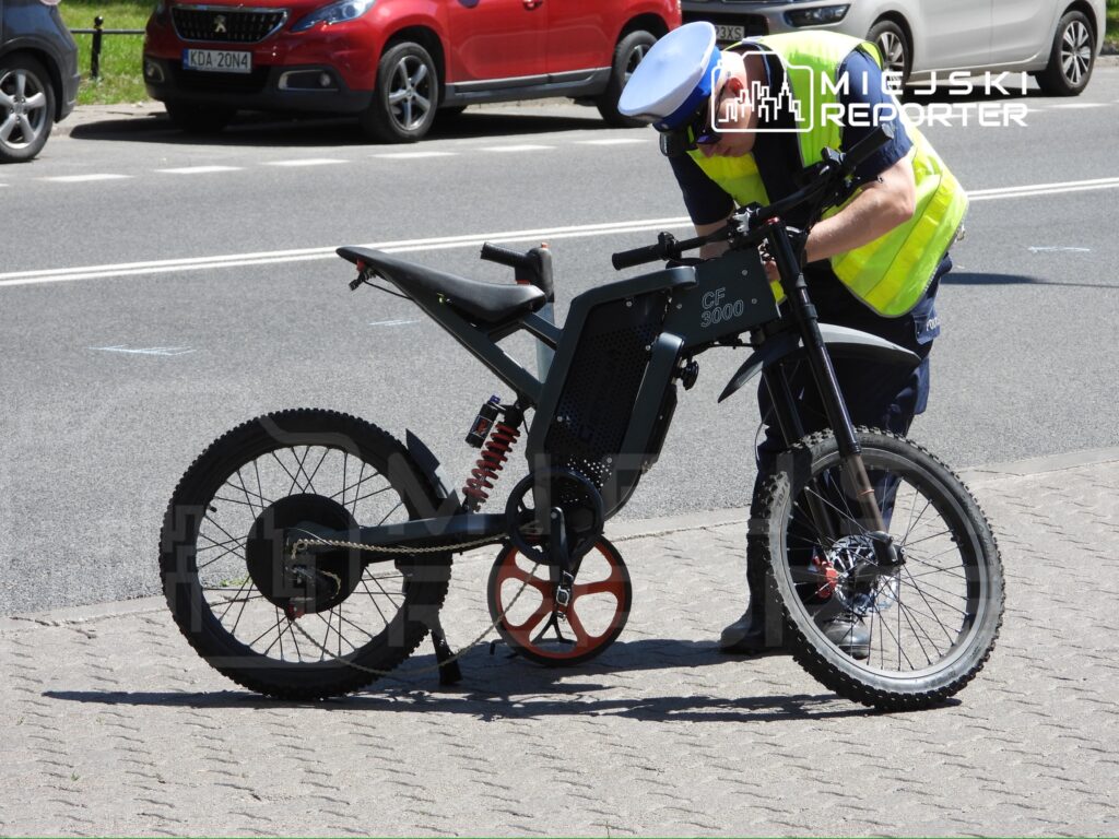 Policjant w kamizelce odblaskowej sprawdza elektryczny rower na chodniku obok ruchliwej ulicy.
