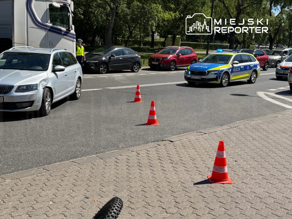 Policyjny radiowóz z włączonymi sygnałami świetlnymi przejeżdża obok stojących samochodów, na drodze ustawione są pachołki.