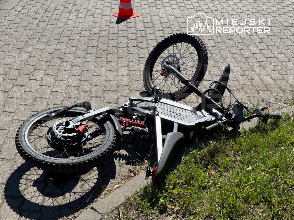 Złamany rower elektryczny leży na chodniku obok trawnika, obok widoczny czerwony pachołek.