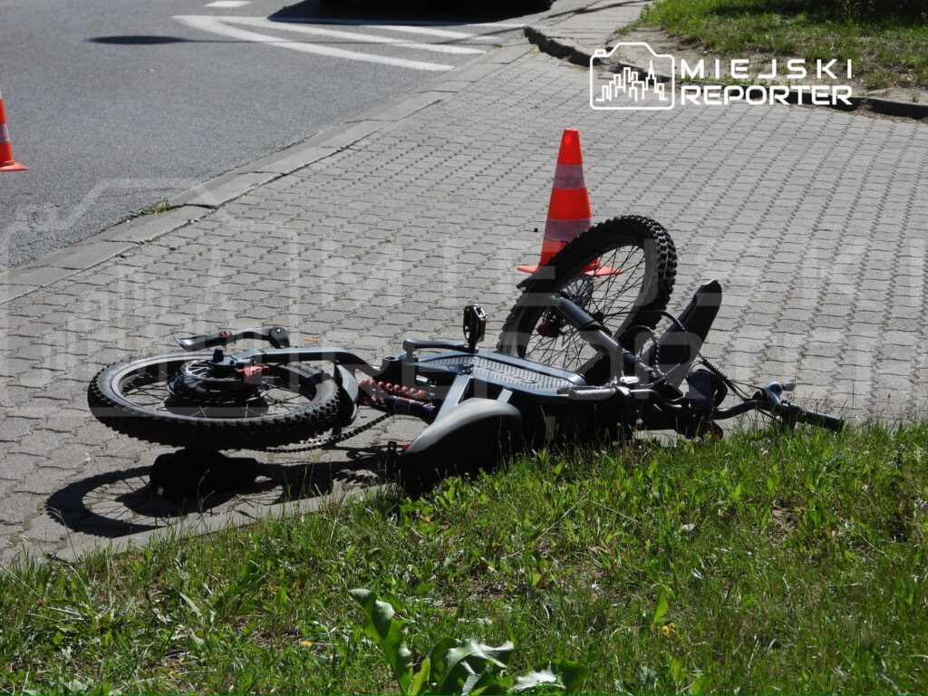 Rozbite rower leży na chodniku obok pomarańczowego pachołka, w tle widać ruchliwą ulicę.