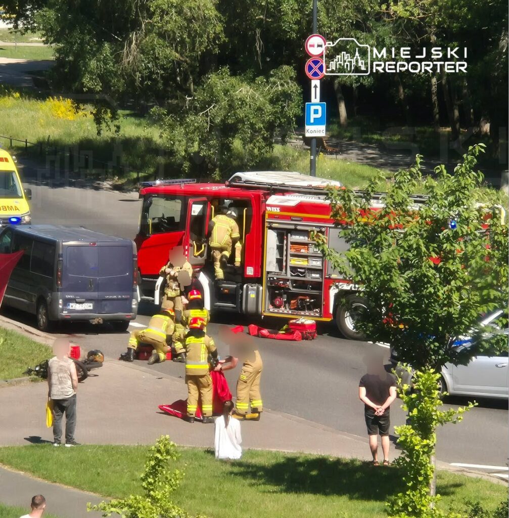 Grupa strażaków w mundurach udziela pomocy poszkodowanemu na jezdni, obok stoi wóz strażacki.
