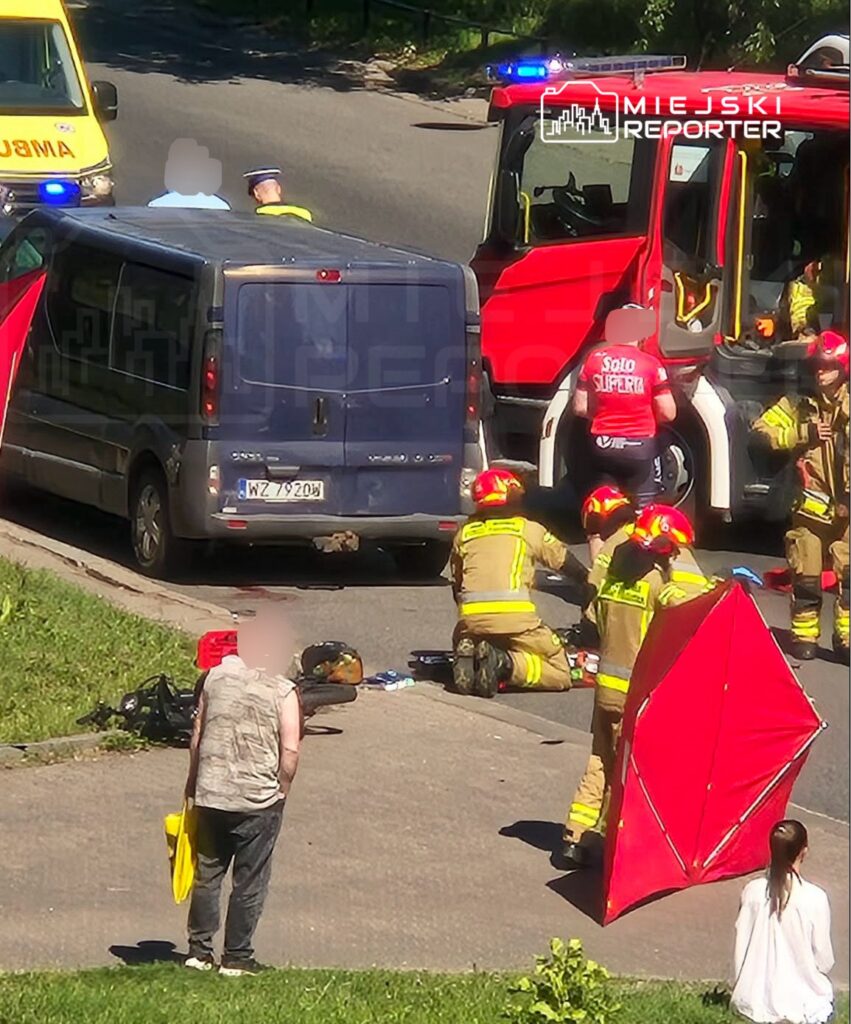 Grupa strażaków udziela pomocy osobie leżącej na drodze, obok zaparkowanego furgonu i wozu strażackiego.