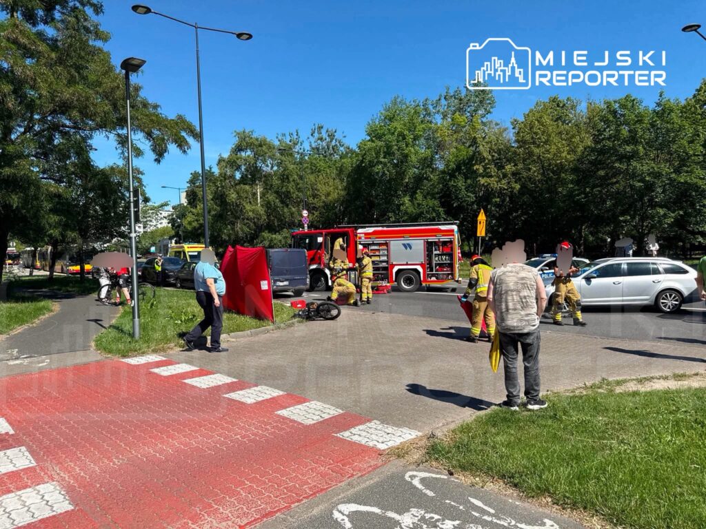 Grupa strażaków i policjantów udziela pomocy na skrzyżowaniu, obok czerwonego wozu strażackiego i zablokowanego ruchu.