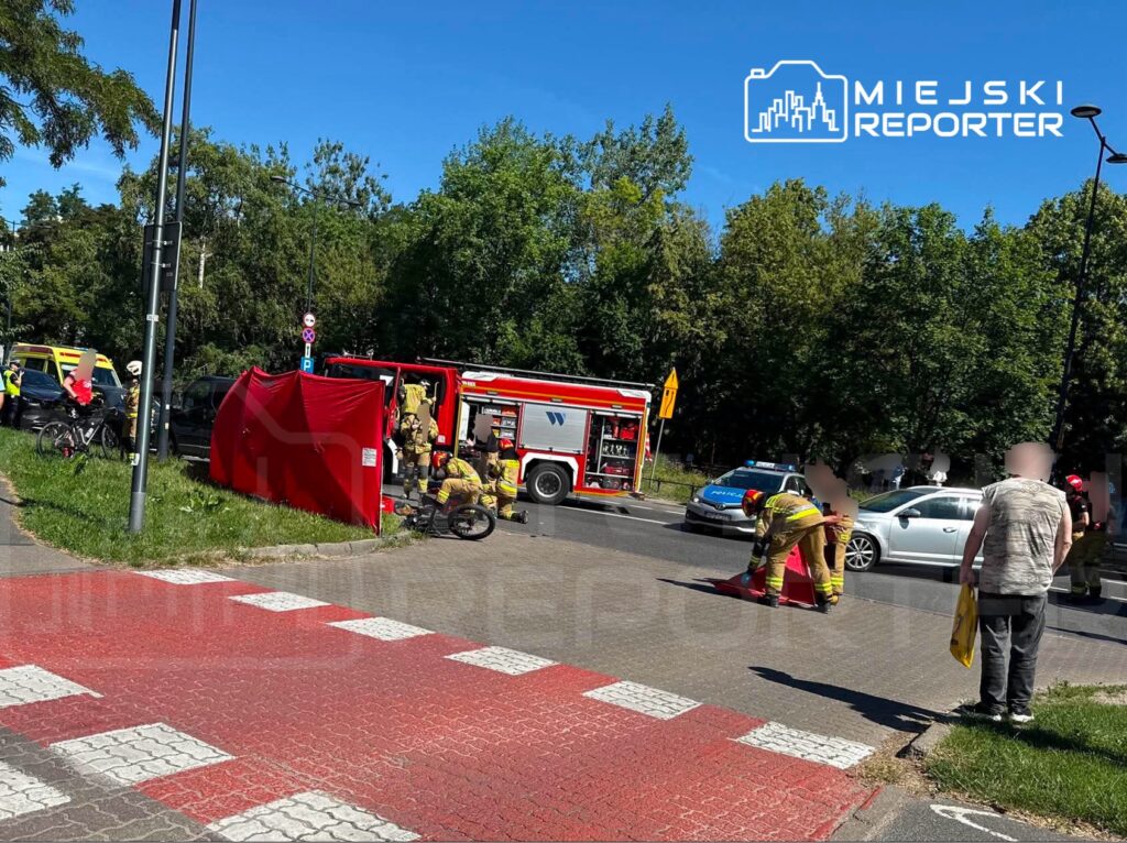 Strażacy zabezpieczają miejsce wypadku drogowego, obok czerwonego wozu strażackiego, na skrzyżowaniu z zielonym otoczeniem.