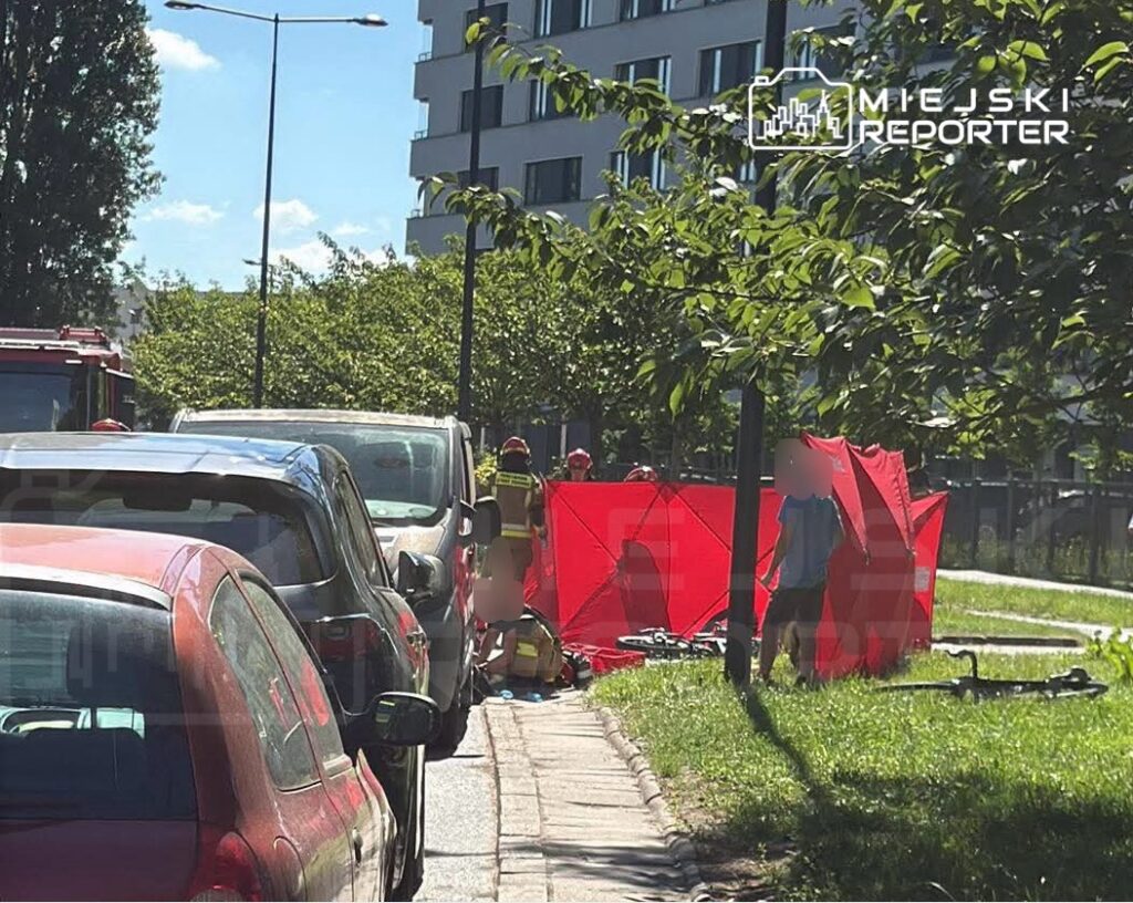 Ratownicy medyczni pracują przy czerwonym namiocie na chodniku, obok zaparkowanych samochodów w miejskim otoczeniu.