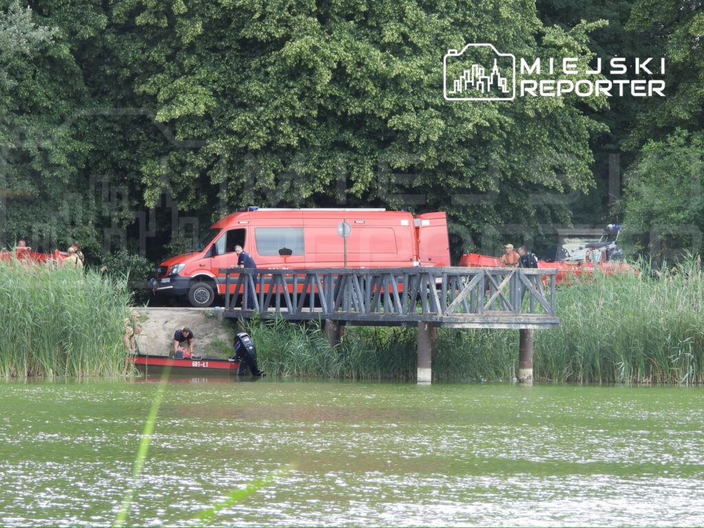 Ratownicy wodni przeszukują brzeg jeziora, obok czerwona furgonetka i łódź motorowa na pomoście.