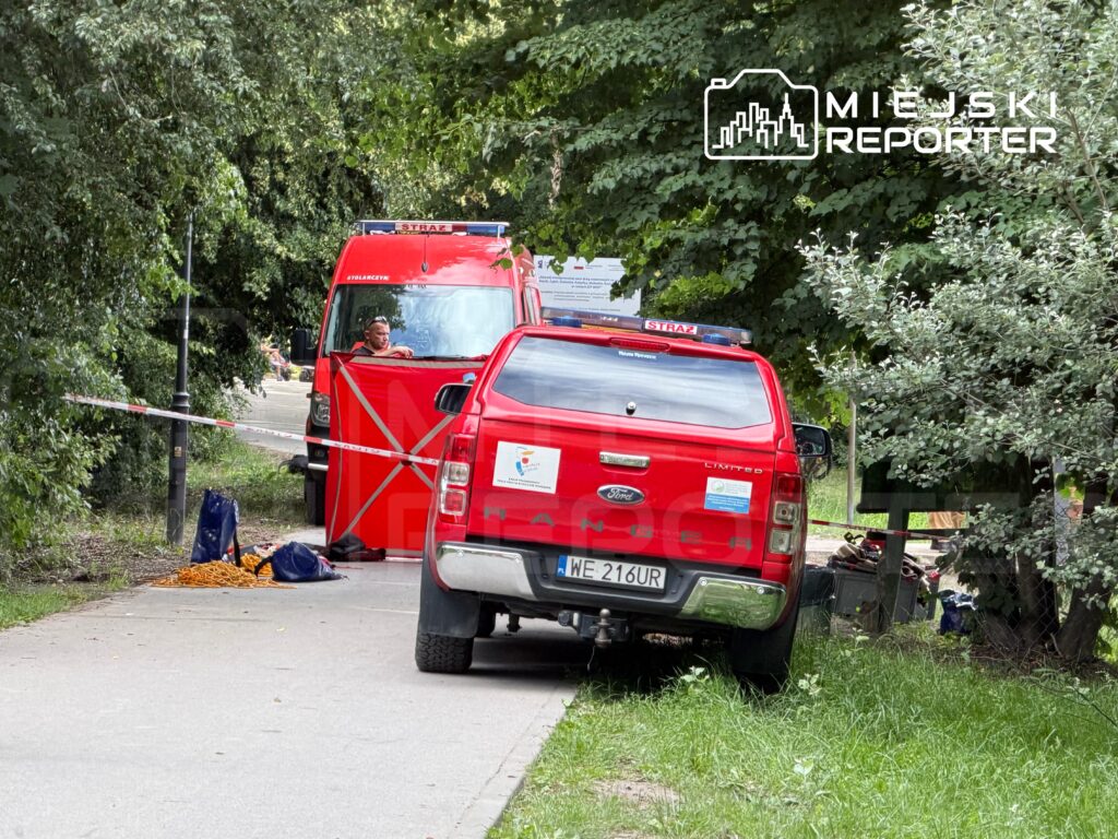 Czerwony samochód strażacki zatrzymany na ścieżce, obok widoczne sprzęty ratunkowe oraz mężczyzna w mundurze strażackim.