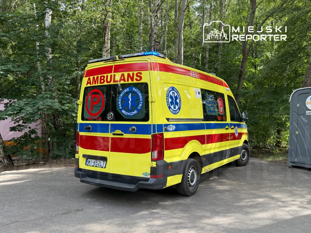 Karetka pogotowia z niebieskimi światłami zaparkowana wśród drzew obok przenośnej toalety.