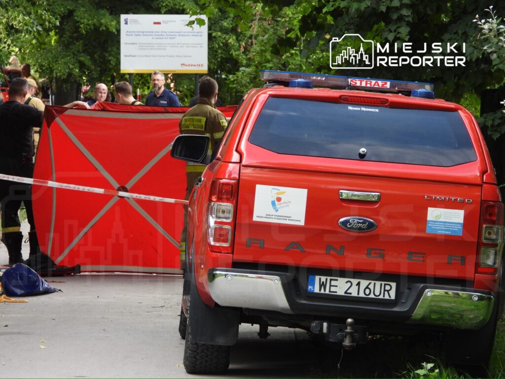 Grupa strażaków i policjantów rozmawia obok czerwonego samochodu ratunkowego, zasłaniając miejsce zdarzenia czerwoną płachtą.