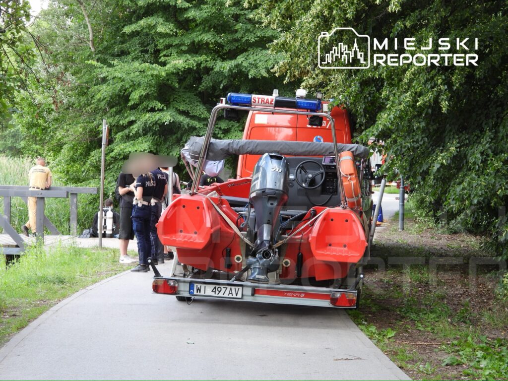 Grupa strażaków i policjantów przy wodnym pojeździe ratunkowym w zaroślach nad brzegiem.