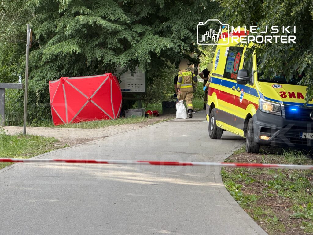 Ratownicy medyczni i strażacy pracują w parku obok czerwonego namiotu, przy zaparkowanej karetce.